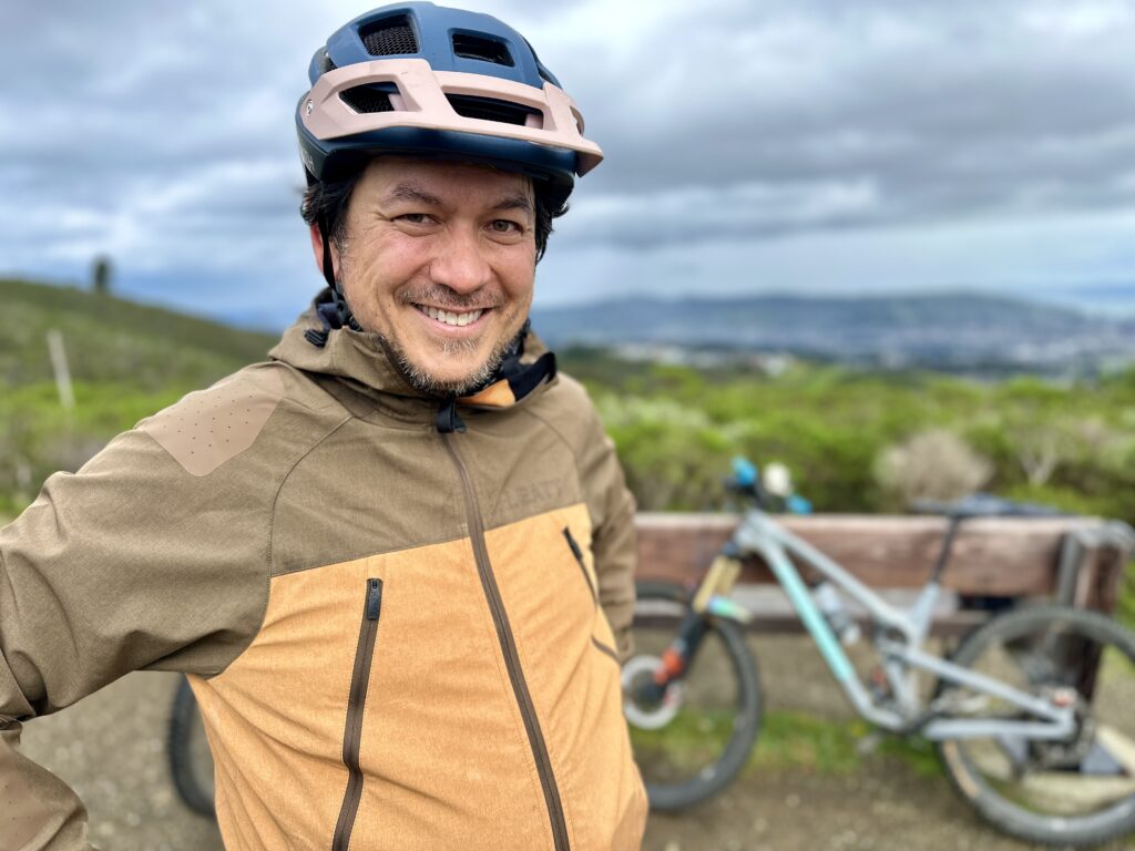 Yonas wears a rain jacket and a bike helmet and smiles at the camera. His bike is out of focus behind him.