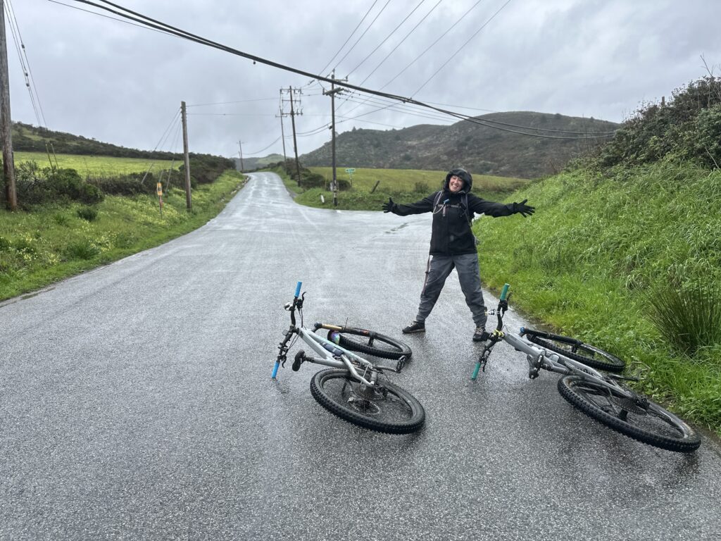 Nicki stands on the side of the road with two bikes at her feet and her arms stretched out. It is a rainy day and the road is wet.