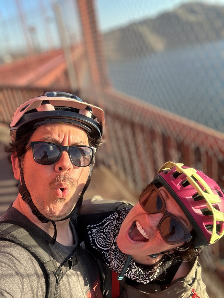 Yonas and Nicki smile at the camera with their helmets on, while standing on the Golden Gate Bridge